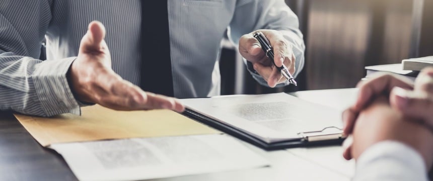 Two people sit across a desk in a corporate law office, one holding a pen and explaining something while pointing to a document on a clipboard.