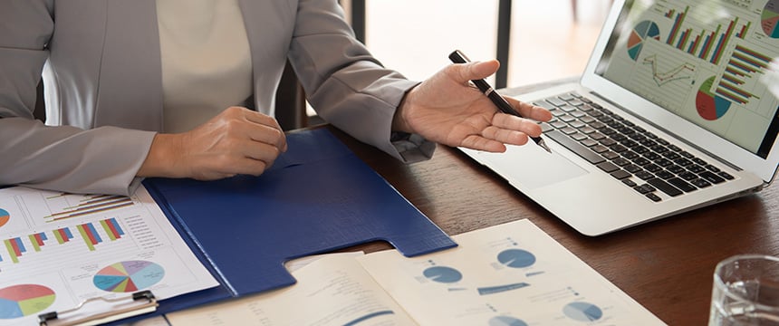 A person in business attire gestures toward a laptop displaying charts and graphs, with printed financial documents spread out on a wooden desk—ideal for chicago lawyers needing litigation support.