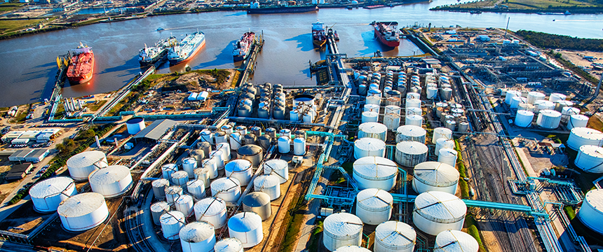 Aerial view of an industrial port with large storage tanks, pipelines, and several cargo ships docked along the river, situated near prominent law offices specializing in corporate law.