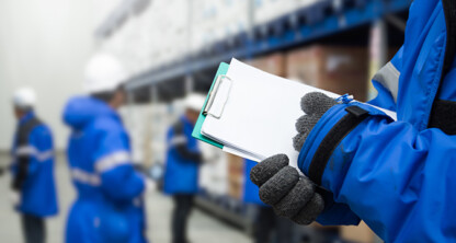 Person in blue jacket and gloves holding a clipboard in a warehouse, while other workers organize shelves of boxes—much like lawyers in Chicago ensuring every detail is accounted for behind the scenes.