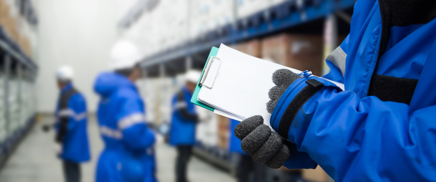 Person in blue jacket and gloves holding a clipboard in a warehouse, while other workers organize shelves of boxes—much like lawyers in Chicago ensuring every detail is accounted for behind the scenes.