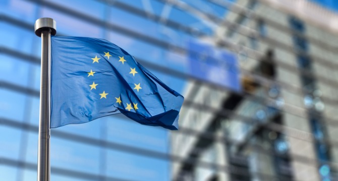 European Union flag with yellow stars on a blue background flying in front of a modern glass building, symbolizing international cooperation at prestigious law offices specializing in corporate law and litigation support.