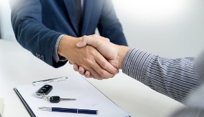 Two people shake hands over a desk with a contract, pen, and car keys, suggesting a business or sales agreement in a corporate law office.
