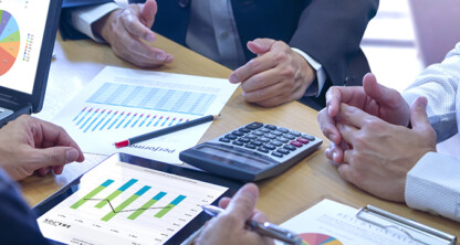 Three people in a corporate law office discuss financial charts at a table with a laptop, tablet, calculator, and printed graphs, focusing on data analysis and business planning.