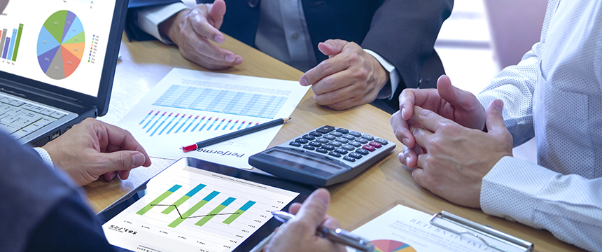 Three people in a corporate law office discuss financial charts at a table with a laptop, tablet, calculator, and printed graphs, focusing on data analysis and business planning.