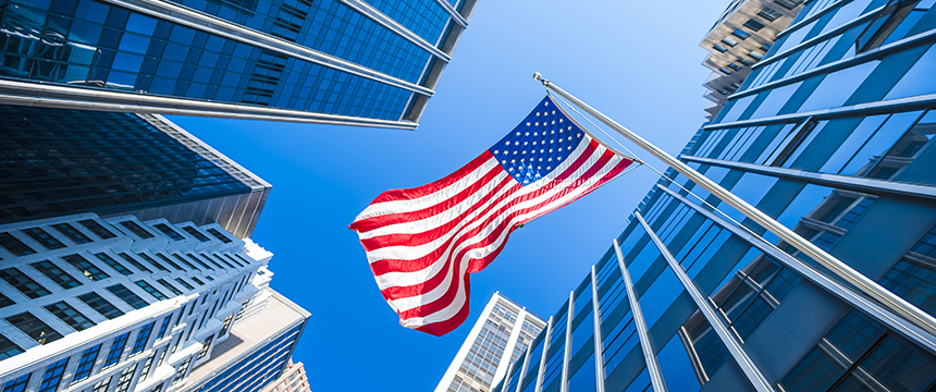 An American flag on a pole is centered among tall modern law offices, viewed from below against a clear blue sky, evoking the strength and integrity of a corporate law office.