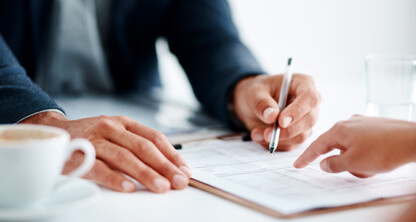 Two people sit at a table, one pointing and the other holding a pen over a document, discussing intellectual property law, with a cup of coffee and a glass of water nearby.