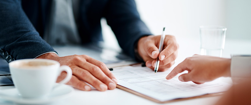 Two people sit at a table, one pointing and the other holding a pen over a document, discussing intellectual property law, with a cup of coffee and a glass of water nearby.