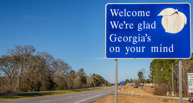 Blue roadside sign reads "Welcome We're glad Georgia's on your mind" with a peach graphic; road and trees visible under clear sky—a friendly sight for travelers, including those from a corporate law office or lawyers in Chicago.
