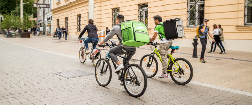 Several cyclists, including food delivery riders with branded backpacks, ride along a city street lined with buildings and pedestrians—an everyday scene where even chicago lawyers might spot potential intellectual property law issues.