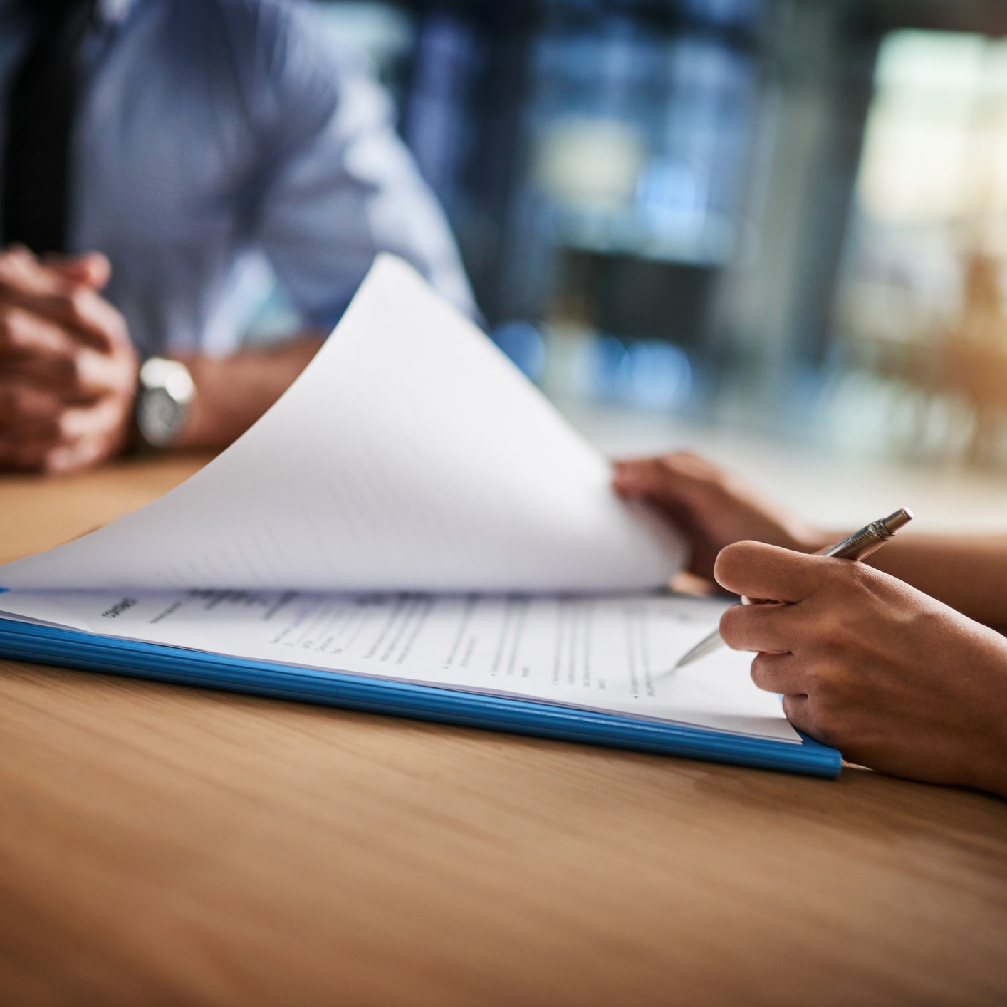 A person holding a pen reviews and flips through documents on a clipboard at a table in a corporate law office, with another individual seated across from them.