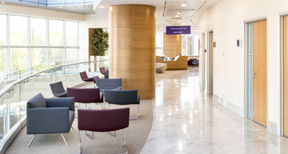Modern hospital waiting area with grey and purple chairs, a curved glass railing, wood-paneled columns, and large windows letting in natural light—ideal for consultations or meetings with lawyers in Chicago. Reception desk visible in the background.