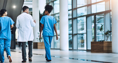 Three healthcare professionals, two in blue scrubs and one in a white coat, walk down a modern hospital corridor with large windows and indoor plants, resembling the sleek atmosphere of a corporate law office.