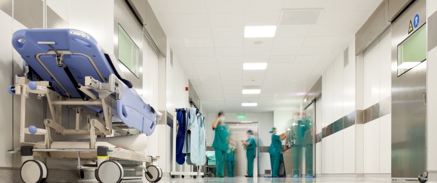 A hospital corridor with a blue gurney on the left and several medical staff in scrubs walking and talking in the background, resembling the bustling energy often seen in a corporate law office.