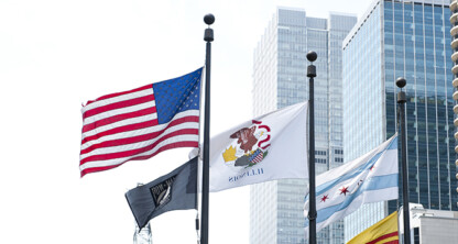 Several flags, including the U.S., Illinois, and Chicago city flags, are flying on poles in front of modern high-rise buildings that house a corporate law office offering litigation support.