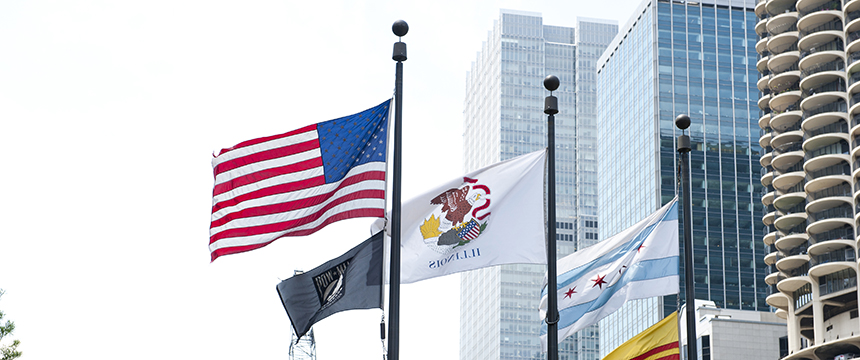 Several flags, including the U.S., Illinois, and Chicago city flags, are flying on poles in front of modern high-rise buildings that house a corporate law office offering litigation support.