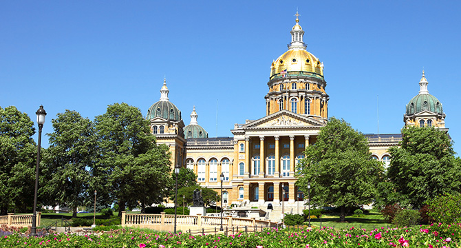 The image shows the Iowa State Capitol building with its gold dome, surrounded by trees and flowers under a clear blue sky, offering a setting that could inspire any corporate law office or provide tranquility amid litigation support work.