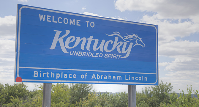 Blue roadside sign reads "Welcome to Kentucky, Unbridled Spirit, Birthplace of Abraham Lincoln," with trees and a partly cloudy sky in the background—a scenic view for travelers, including chicago lawyers or those seeking litigation support.