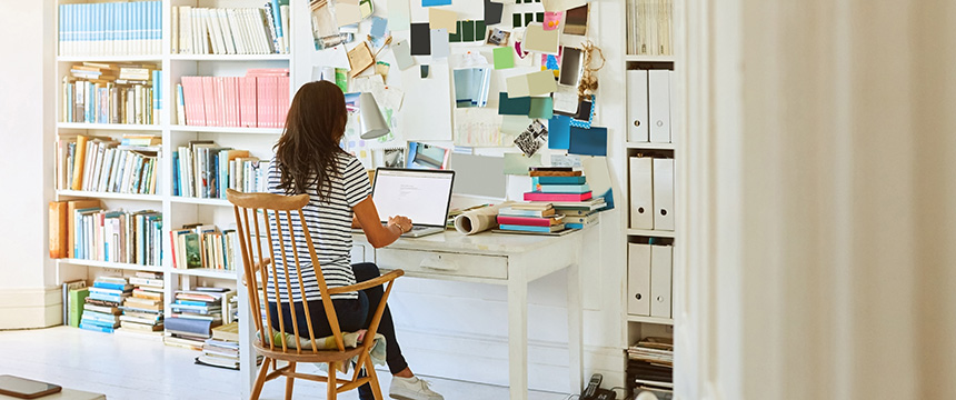 A woman sits at a desk working on a laptop in a home office, surrounded by bookshelves, binders, stacks of books, and a bulletin board—her workspace resembles the busy law offices of lawyers in Chicago.