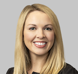 A woman with straight blonde hair, wearing a black top, smiles at the camera against a plain gray background, reflecting the professionalism often seen in corporate law offices.