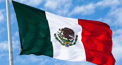 The flag of Mexico with green, white, and red vertical stripes and a national emblem in the center is shown flying against a partly cloudy sky, symbolizing pride often seen outside law offices or among Chicago lawyers.