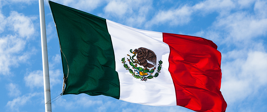 The flag of Mexico with green, white, and red vertical stripes and a national emblem in the center is shown flying against a partly cloudy sky, symbolizing pride often seen outside law offices or among Chicago lawyers.