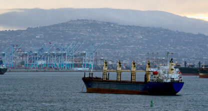 A cargo ship is anchored in a harbor near cranes and shipping containers, with hills rising under a cloudy sky—a busy scene often handled by law offices specializing in maritime contracts and litigation support.