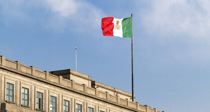 A Mexican flag flies atop a beige government building under a blue sky with some clouds, reminiscent of the professional presence found at top corporate law offices.