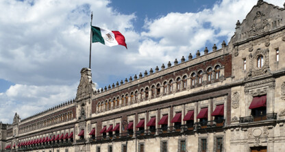 The Mexican flag flies above the National Palace, a historic government building with ornate architectural details and red awnings, once housing important law offices, under a partly cloudy sky.