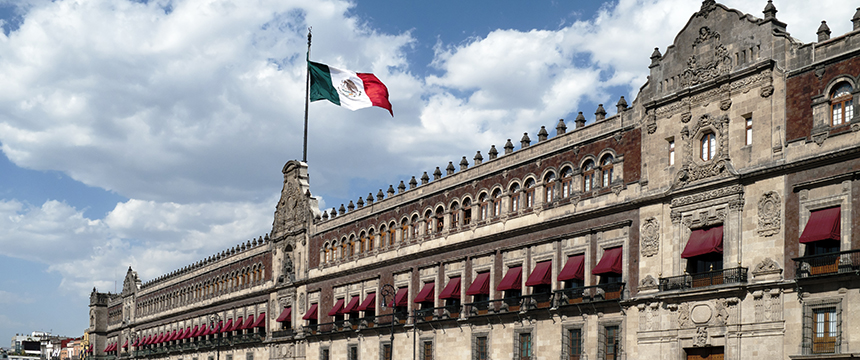 The Mexican flag flies above the National Palace, a historic government building with ornate architectural details and red awnings, once housing important law offices, under a partly cloudy sky.