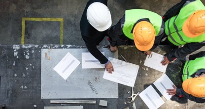 Four workers wearing safety helmets and reflective vests review blueprints and documents—potentially related to intellectual property law or litigation support—on a worktable in an industrial setting, viewed from above.