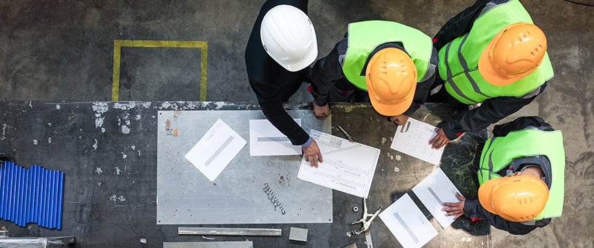 Four workers wearing safety helmets and reflective vests review blueprints and documents—potentially related to intellectual property law or litigation support—on a worktable in an industrial setting, viewed from above.