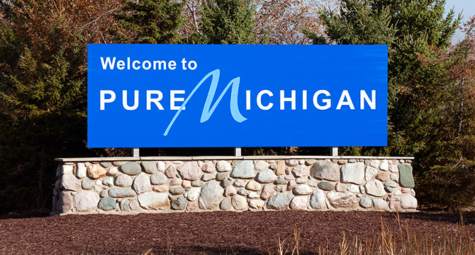 A blue roadside sign reads "Welcome to Pure Michigan" in white letters, mounted on a stone base with trees in the background—a scene familiar to many Chicago lawyers traveling for litigation support.