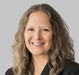 A woman with long, wavy gray hair wearing a black blazer smiles in front of a plain light gray background, reflecting the professionalism of lawyers in Chicago and their expertise in intellectual property law.