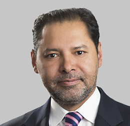Man with short dark hair and beard, wearing a dark suit, white shirt, and striped tie, posed in front of a plain gray background—typical of professional portraits for lawyers in Chicago.