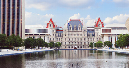 The image shows the New York State Capitol building in Albany, reflected in a large rectangular pool, surrounded by trees and modern buildings—an inspiring scene for law offices or firms offering litigation support.