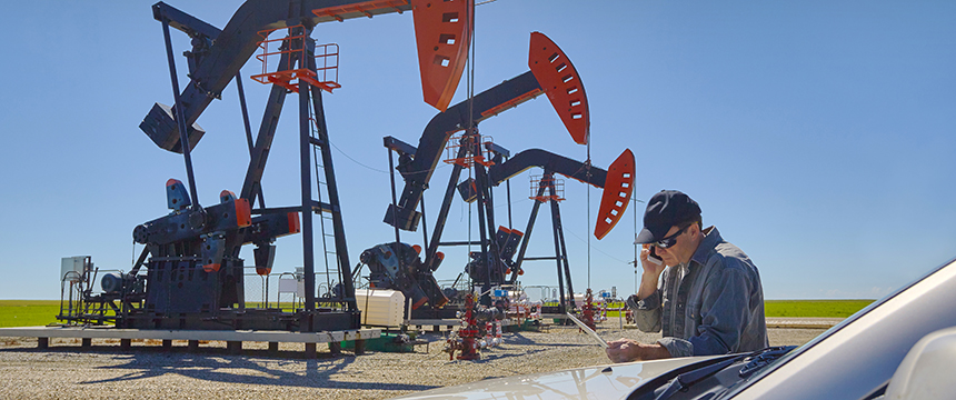 A man in a cap looks at papers and talks on the phone near several oil pumpjacks, possibly discussing litigation support, under a clear blue sky.