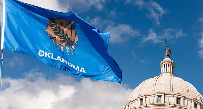 The Oklahoma state flag waves in front of the dome of the Oklahoma State Capitol building under a partly cloudy sky, reminiscent of scenes outside corporate law office buildings across the country.