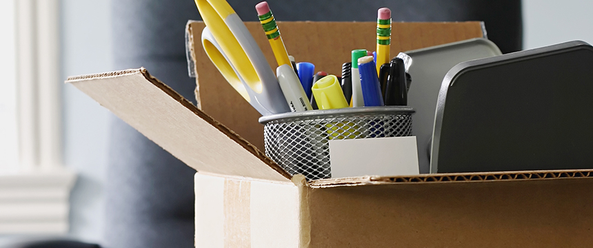 Open cardboard box filled with office supplies, including pens, pencils, scissors, highlighters, a metal cup, and small trays—ideal for a corporate law office—positioned on a desk with a blurred background.