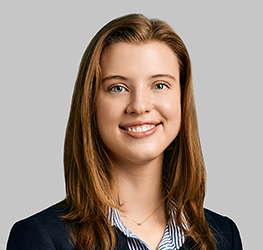 A young woman with straight light brown hair, wearing a dark blazer over a striped shirt, is smiling at the camera against a plain grey background, representing litigation support for lawyers in Chicago.