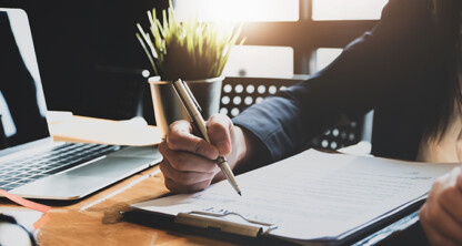 Person in business attire writing on a document with a pen at a desk, with a laptop, plant, and scales of justice visible—a typical scene in corporate law offices or among lawyers in Chicago.