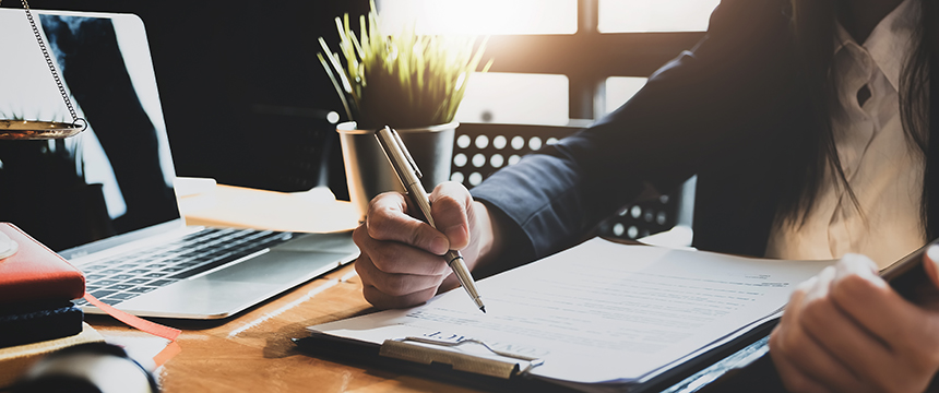 Person in business attire writing on a document with a pen at a desk, with a laptop, plant, and scales of justice visible—a typical scene in corporate law offices or among lawyers in Chicago.