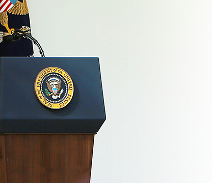 A podium with the Seal of the President of the United States stands outside near a glass door, with an American flag and eagle emblem in the background—reminiscent of a formal address at a prestigious corporate law office.