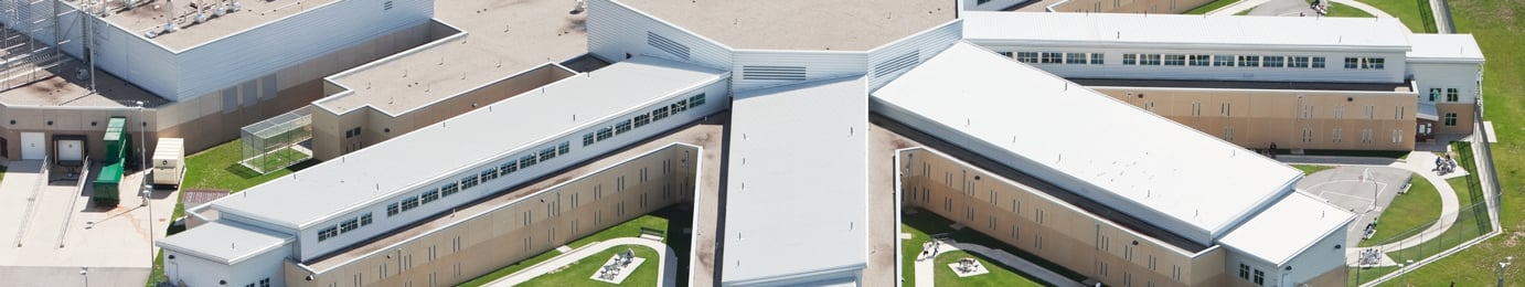 Aerial view of a large, modern correctional facility with multiple connected buildings and fenced outdoor areas, situated near corporate law offices.