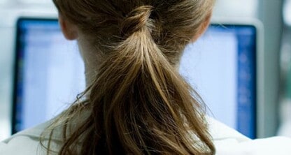 A person with brown hair tied in a ponytail sits facing a computer monitor in a bright indoor setting, typical of modern law offices.