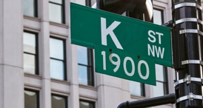 Green street sign reads "K St NW 1900" against a backdrop of a modern office building with large windows, evoking the professionalism of a corporate law office.