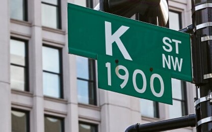 Green street sign reads "K St NW 1900" against a backdrop of a modern office building with large windows, evoking the professionalism of a corporate law office.