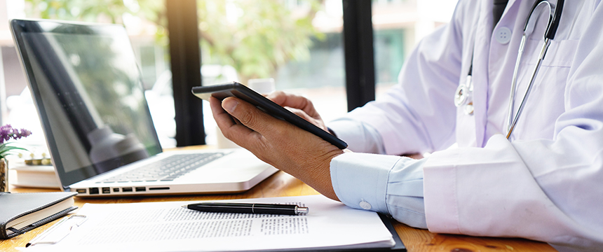 A doctor in a white coat uses a tablet at a desk with a laptop, documents, and a pen, providing litigation support for law offices.