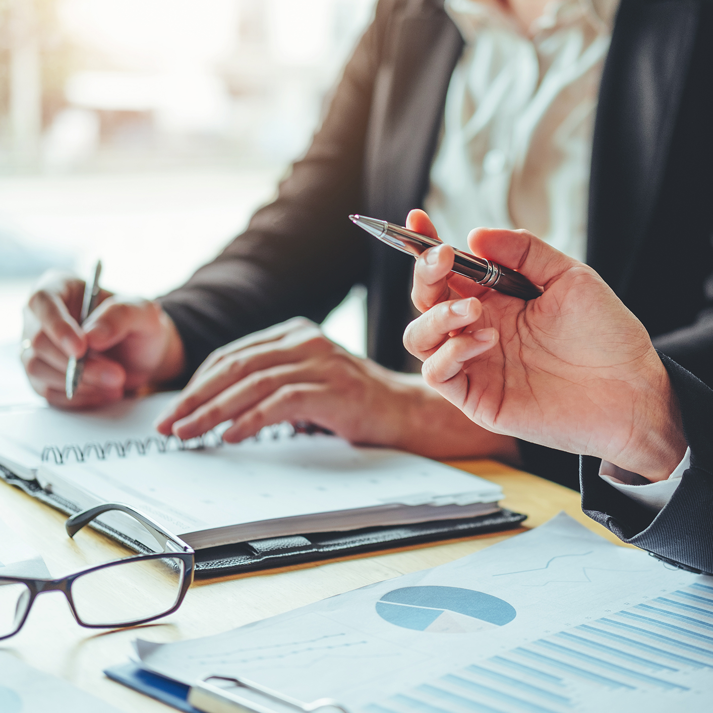 Two people in business attire are seated at a desk in a corporate law office, writing in notebooks with charts and glasses on the table in front of them.