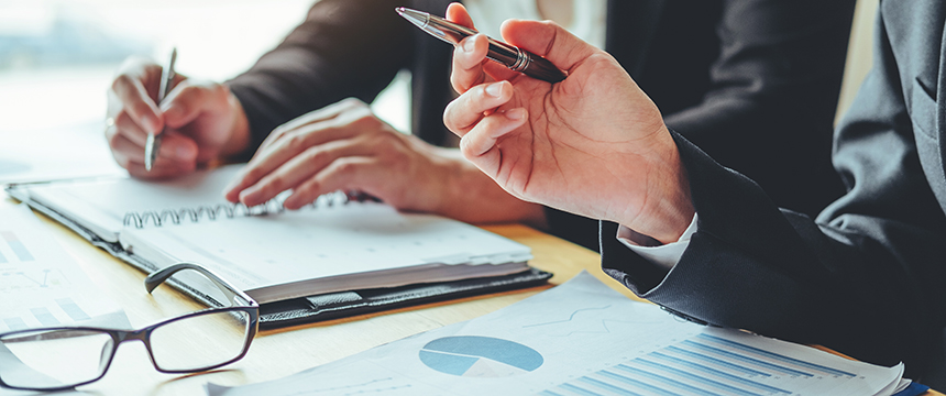 Two chicago lawyers in business attire review and discuss documents and charts at a desk, offering litigation support, with one holding a pen and notebook while eyeglasses rest on the table.